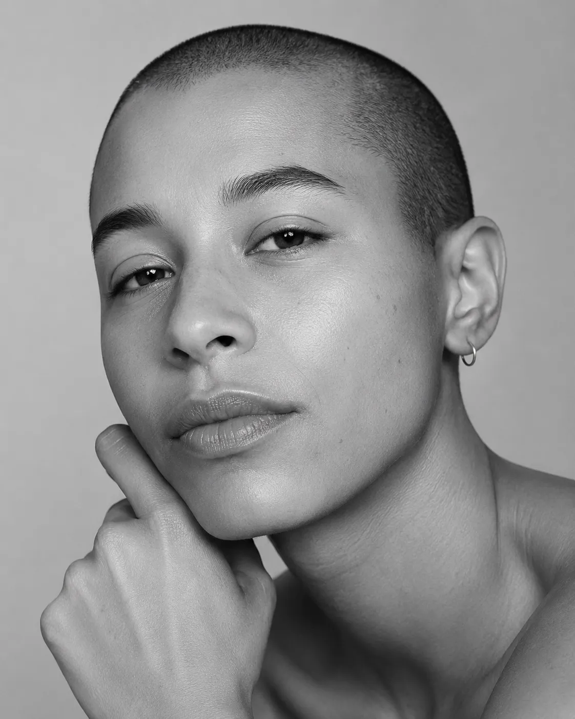 Black and white studio portrait of a person with a closely shaved head, hand resting near the chin